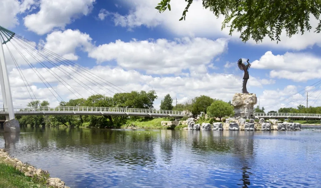 The Keeper of the Plains statue on its rock promontory where the Arkansas rivers meet in Wichita, with the cable-stayed pedestrian bridge crossing the water beside it.