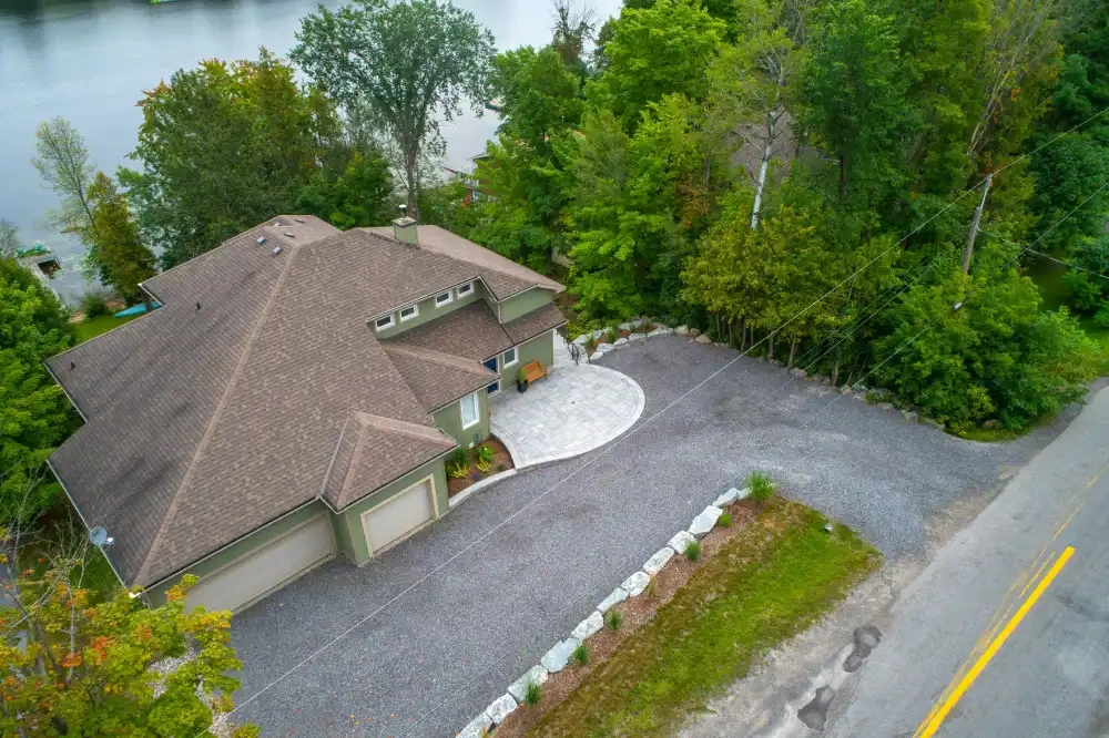 A lakeside house from the air: a paver entry court, a long gravel drive edged with boulders and new planting, and woods either side.