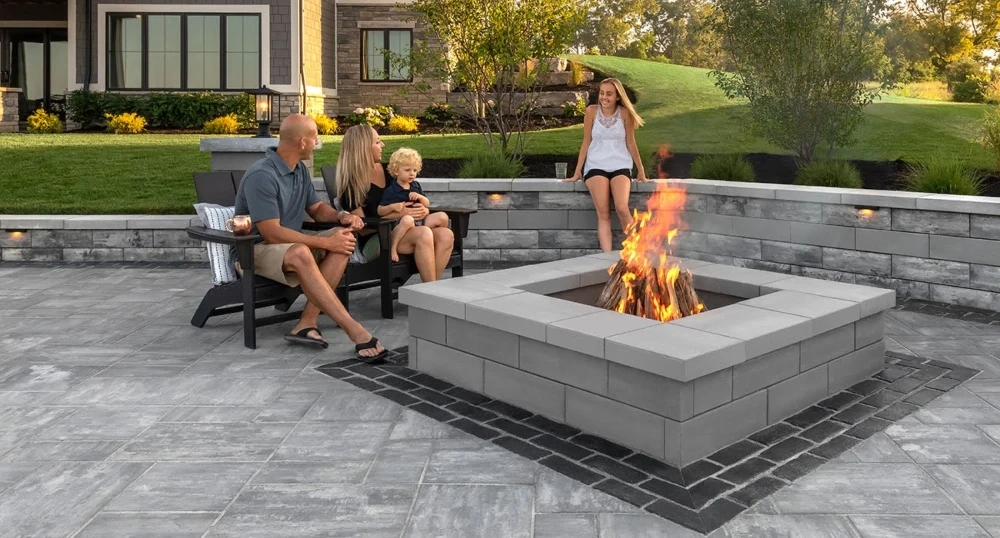 A family seated around a square block fire pit burning on a stamped concrete patio, a low block seat wall and lit house behind them at golden hour.