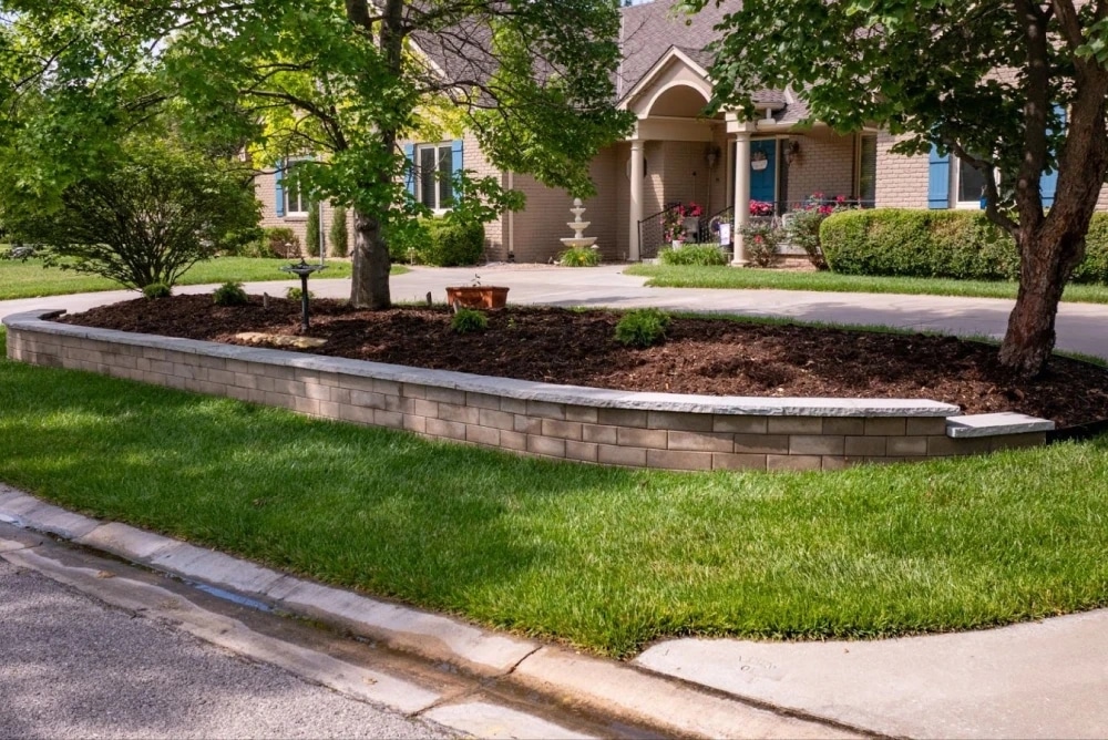 A curved tan block wall with a smooth cap raising a mulched corner bed under two trees, mown lawn and a street corner in front, a stone house behind.