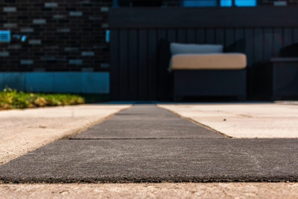 A dark paver walk strip at ground level, running between pale concrete panels toward a shaded seating corner.
