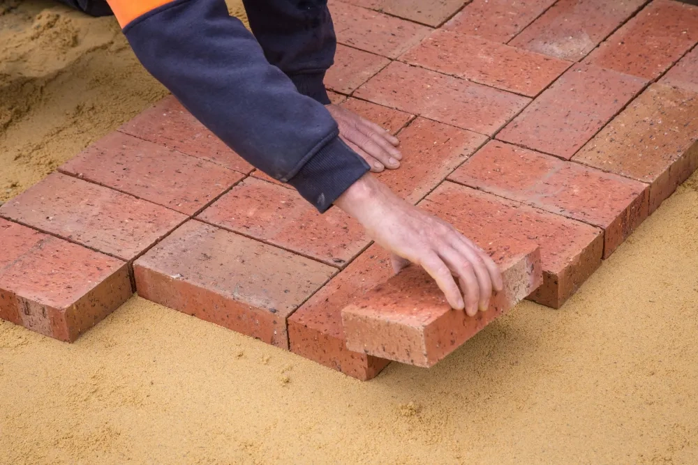 Two hands setting red clay pavers into a screeded sand bed, the laid field running away to one side.