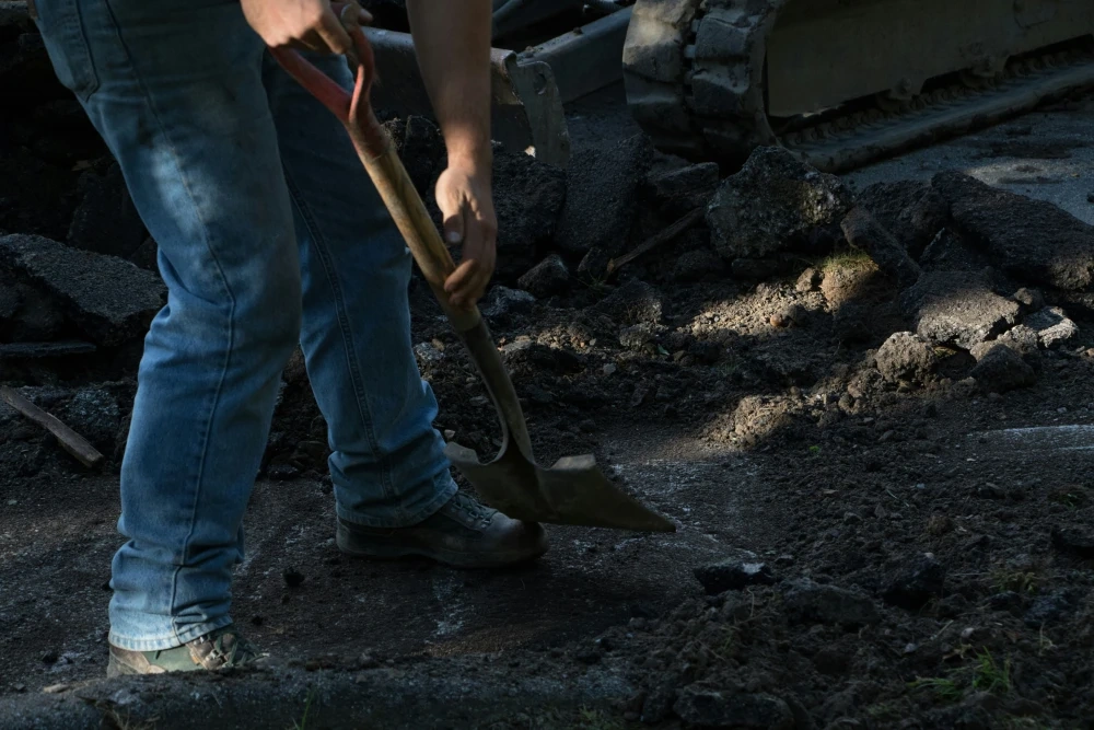 A worker in jeans driving a spade into broken dark ground, the tracks of a compact machine behind.