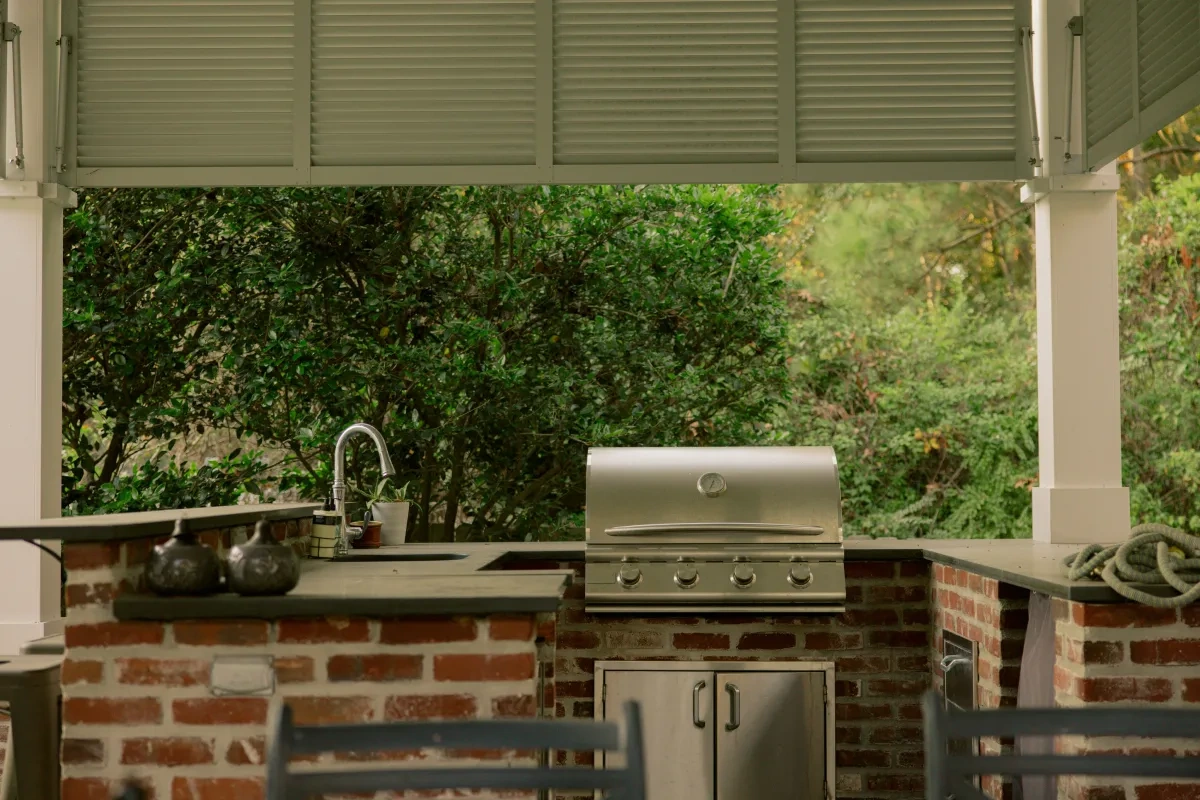 A brick outdoor kitchen under a louvered pavilion roof: built-in grill, dark stone counters and a sink, dense green planting beyond.