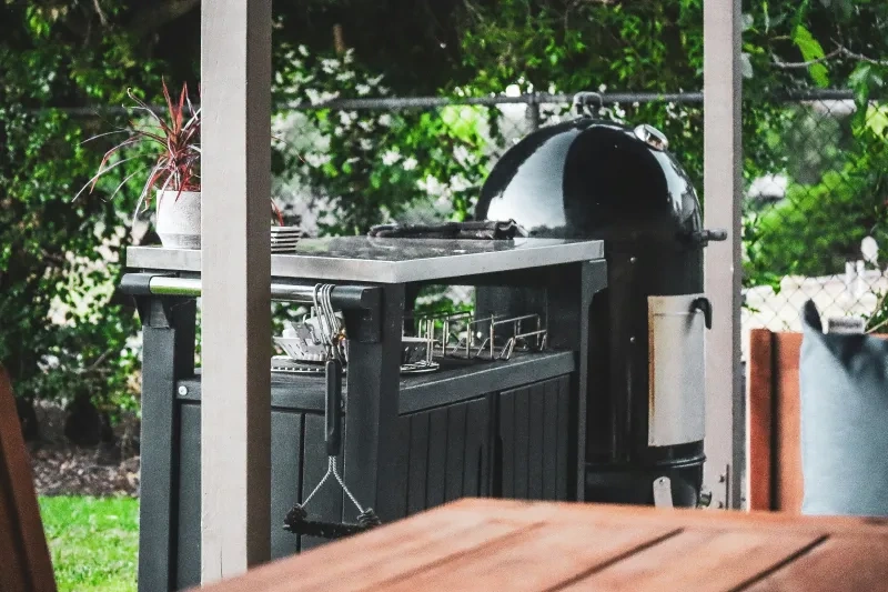 A black kettle smoker beside a stainless-topped prep cart with hanging tools, under a pergola with a fence and shrubs behind.