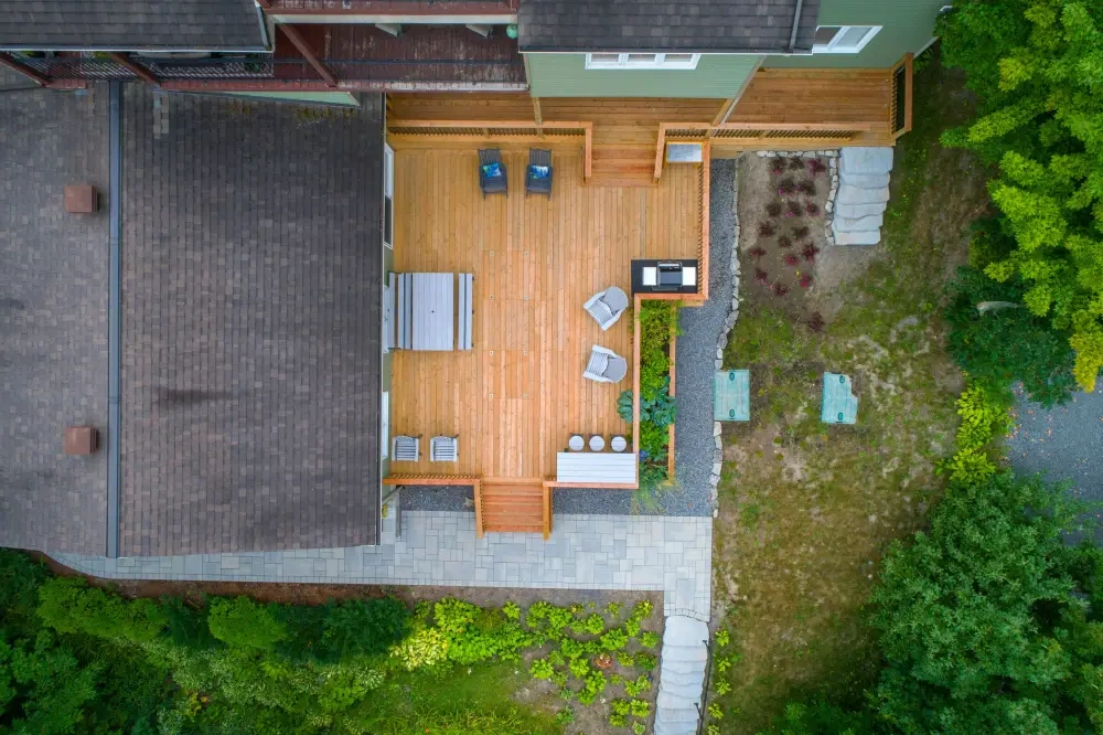 A cedar deck from directly above: loungers and a grill on the boards, a paver path alongside, and a planted slope with a stone retaining edge.