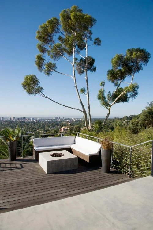 A hillside deck with a white-cushioned corner sofa and concrete fire table, eucalyptus above and a city skyline far below.