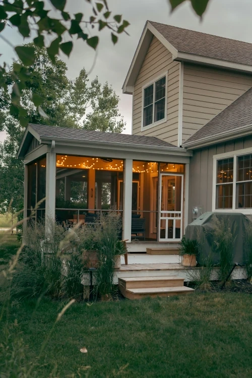 A screened porch glowing with string lights at dusk, wooden steps down to a lawn and tall grasses either side.