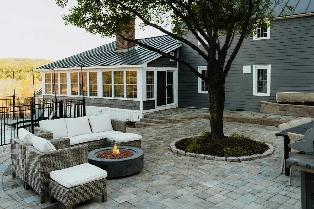 A grey paver patio with wicker seating around a round gas fire bowl, a tree in a stone-ringed bed at its centre and a glassed porch behind.