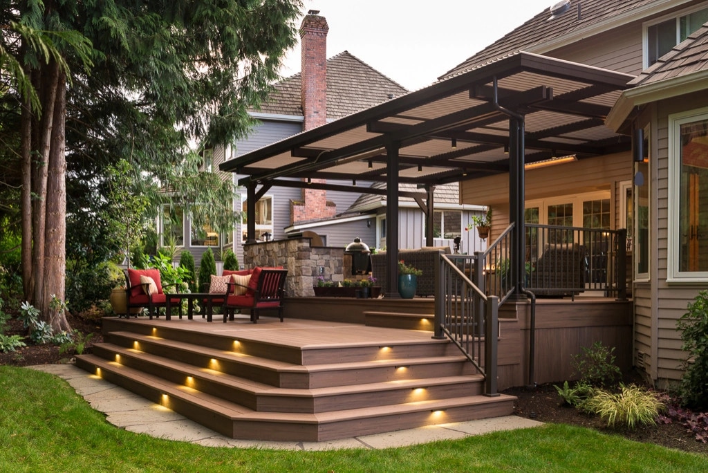 A covered composite deck at dusk: red-cushioned chairs under a slatted roof, a stone outdoor kitchen behind, and riser lights washing the curved steps.