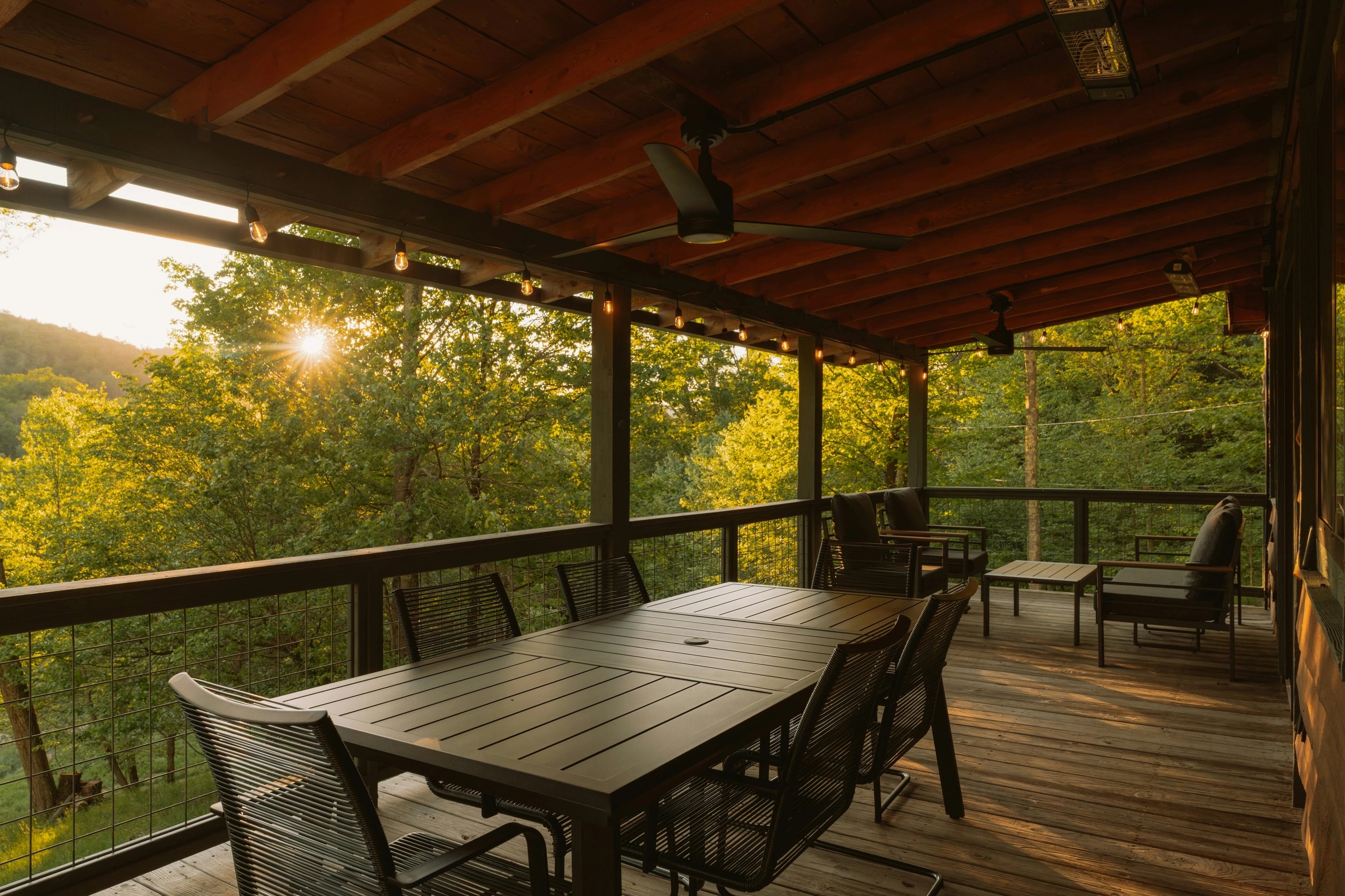 A covered deck under a solid wood roof with ceiling fans, string lights and patio heaters above a dining table, late sun in the trees beyond.