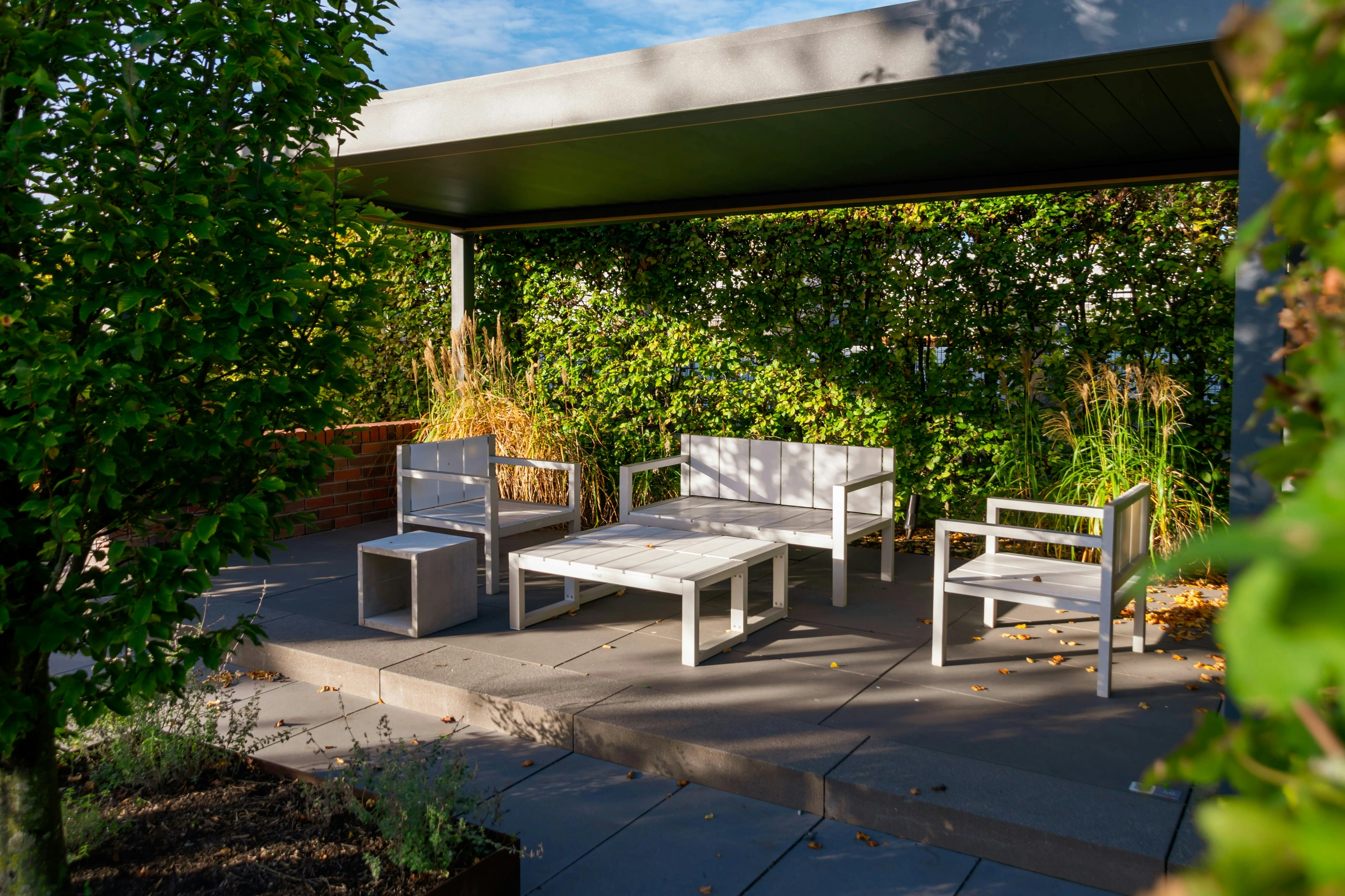 A flat-roofed pavilion over a large-format paver patio with an outdoor sofa and chairs, ornamental grasses and a green hedge behind it.