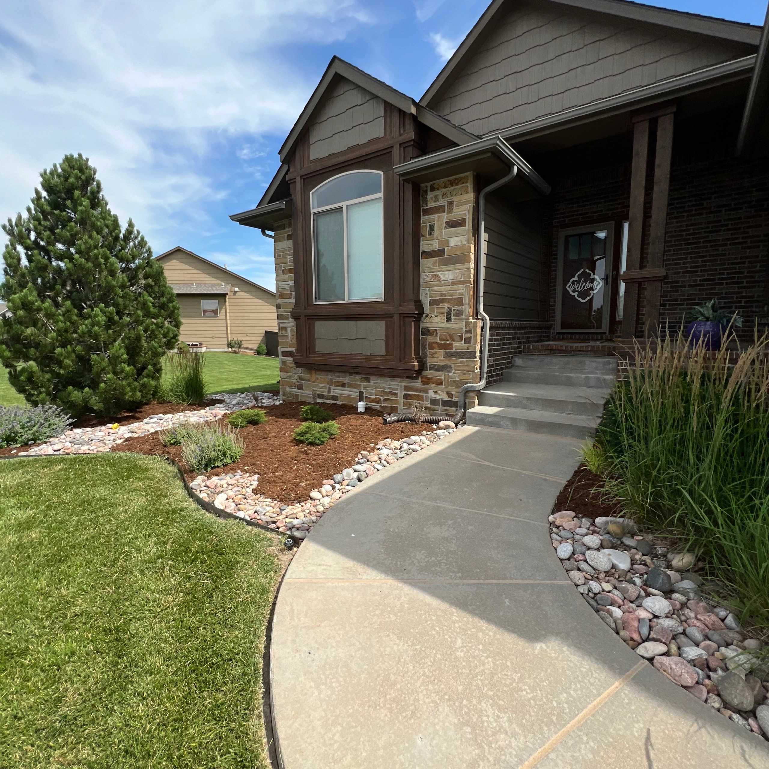 A curved concrete walk to a stone-faced front entry, between new beds of river rock and mulch planted with ornamental grasses, young shrubs and a pine.