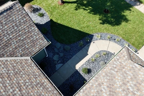 A concrete walk curving between the house and its garage from above, with black slate rock beds and young grasses along it.