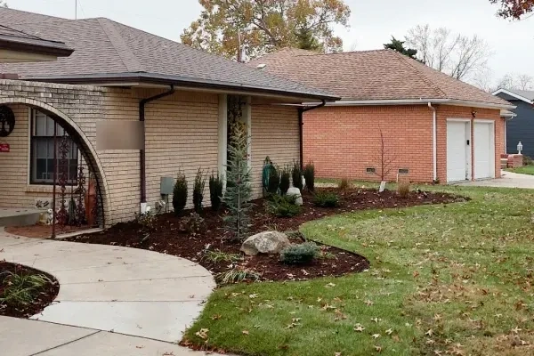 A brick frontage with new columnar junipers, a young spruce and a boulder set in a fresh mulched bed along the curved walk.