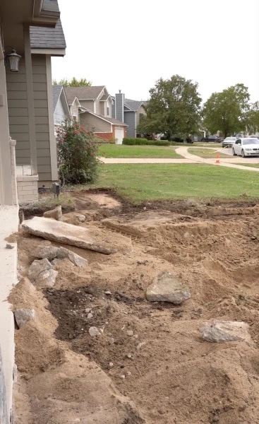 A front garden stripped back to bare soil with stone staged across it ready to be set, the suburban street and neighbouring houses in the background.