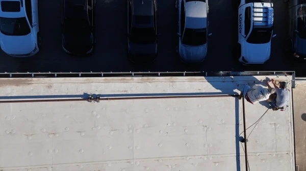 Looking straight down from above onto two crew members working along a commercial roofline, with the parked cars of the lot below them.