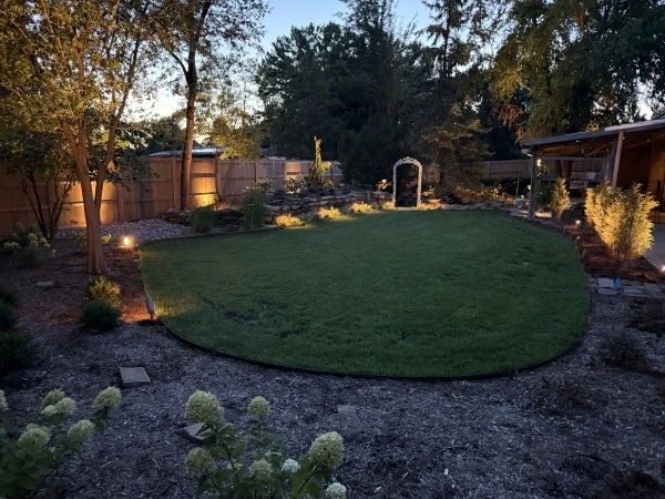A back garden at dusk, with uplights washing the trees along a cedar fence, path lights through the beds and a lit arbour at the far end of the lawn.