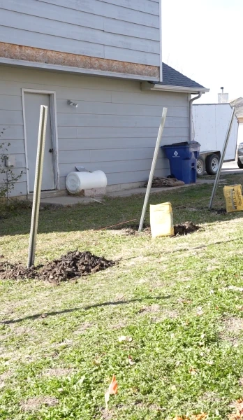 Fence posts braced plumb in freshly dug holes along the side of a house, with bags of concrete mix waiting on the grass beside them.