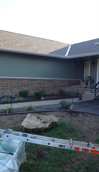 A job site at dusk: a planted bed with path lights already set along a house, a large limestone boulder waiting on the lawn, and a ladder laid down beside it.