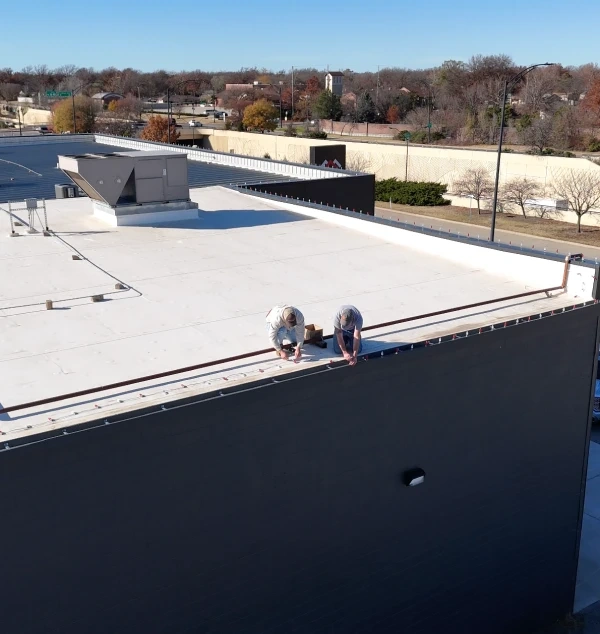 Two crew members kneeling along the parapet of a flat commercial roof, fixing a run of trim, with the town and its bare autumn trees behind them.