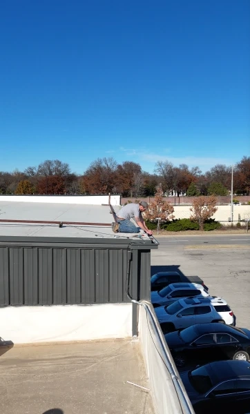 A crew member working along the edge of a commercial roof above a line of parked cars, under a clear winter sky.