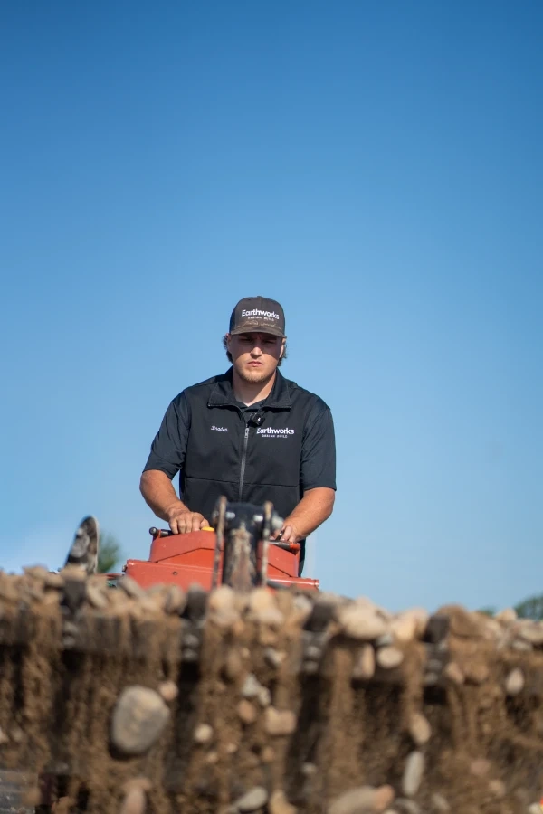 A crew member in Earthworks cap and vest working a machine across a bed of river rock, eyes down, under a clear summer sky.