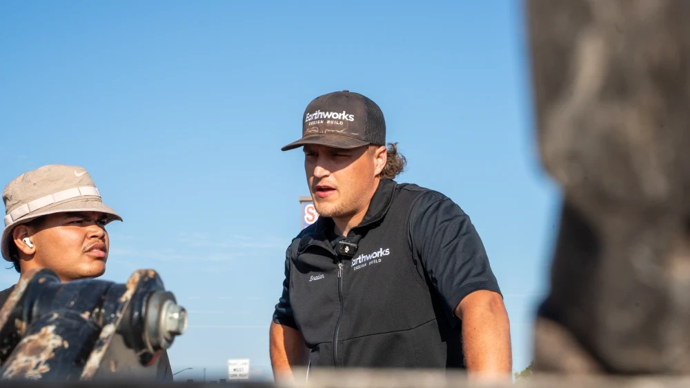 Two Earthworks crew members in branded workwear talking across the arm of an excavator on a job site, under a clear sky.