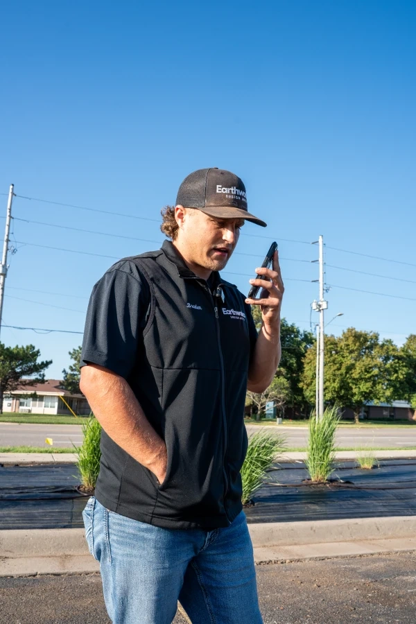 A crew member in Earthworks cap and vest taking a phone call at the kerb, with new ornamental grasses planted through fabric in the bed behind him.