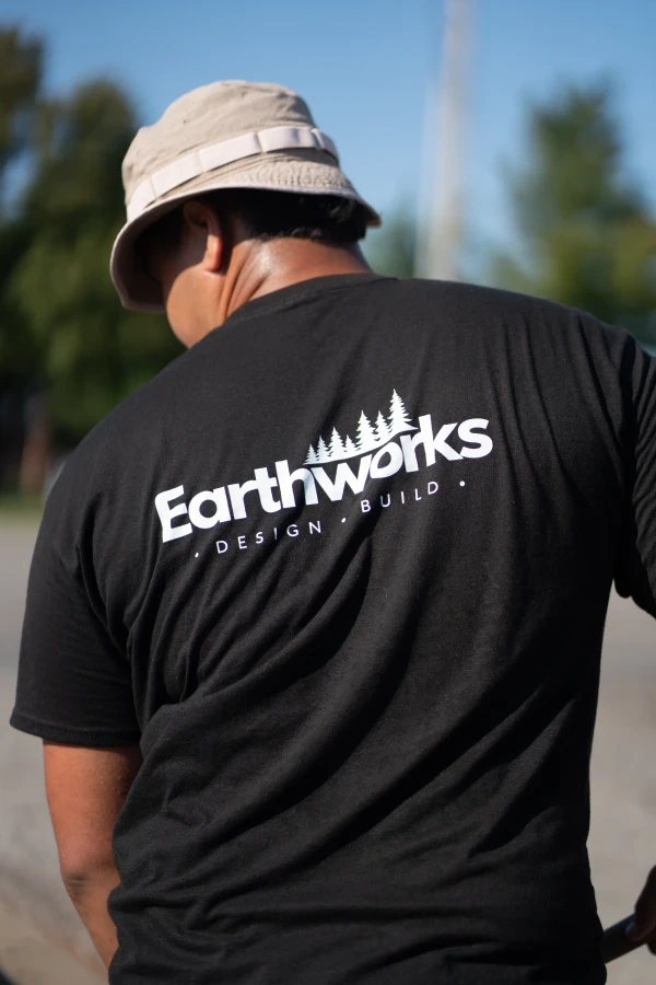 The back of an Earthworks Design·Build work shirt, worn mid-job in a sunlit yard, bucket hat above and gravel below.