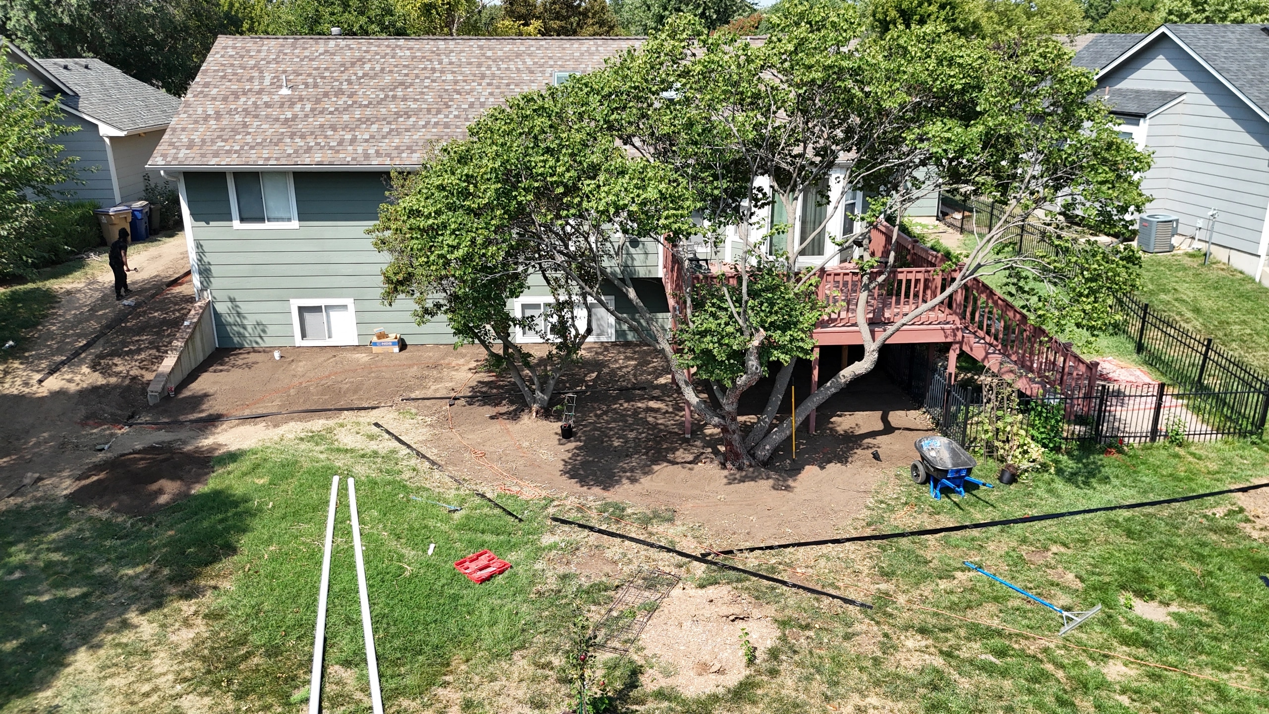 A back yard from above mid-build: the ground around the house and a mature tree stripped to bare soil, with edging and pipe laid out on the lawn.
