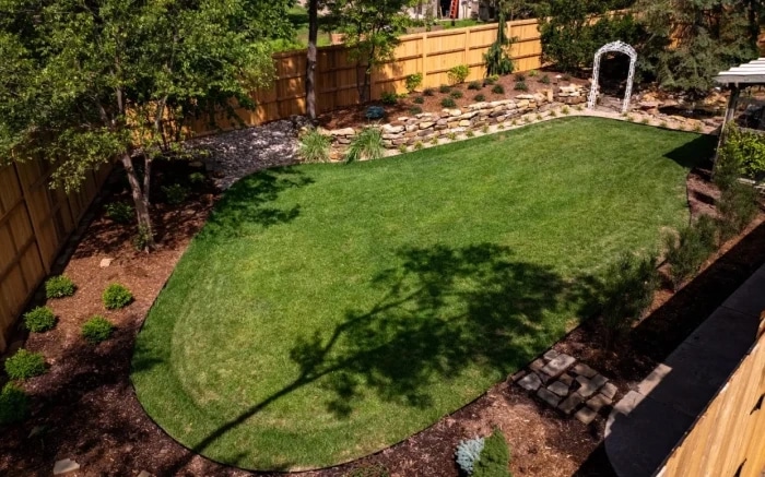A finished back garden from above: a shaped lawn ringed by mulched beds, a low stone wall along the back boundary and a white arbour against cedar fencing.