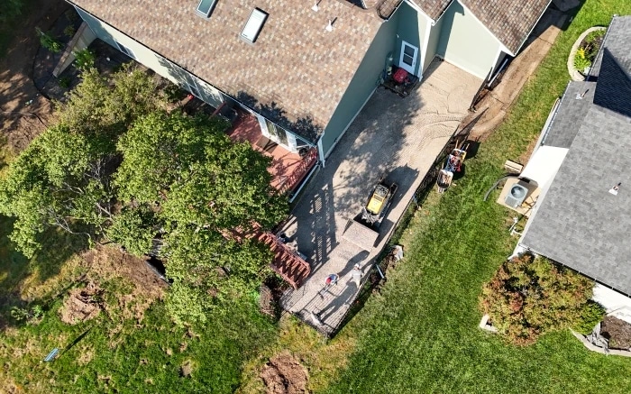 Looking straight down on a compacted gravel base laid between two houses, a roller parked mid-surface and the crew working at its edge.