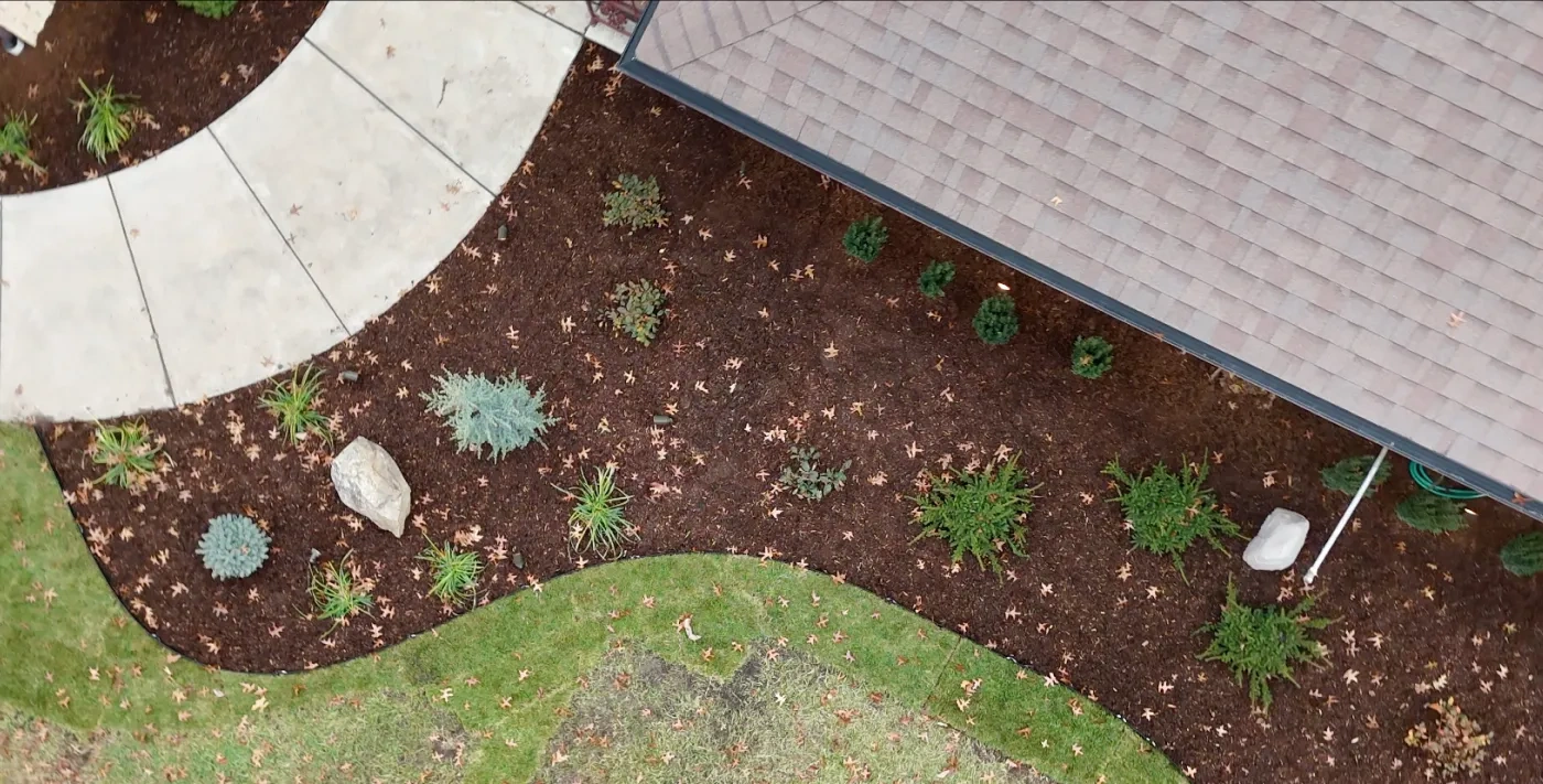Looking straight down on a curved cut-stone patio edge meeting a freshly mulched bed of new shrubs, grasses and set boulders, with the lawn curving away below.