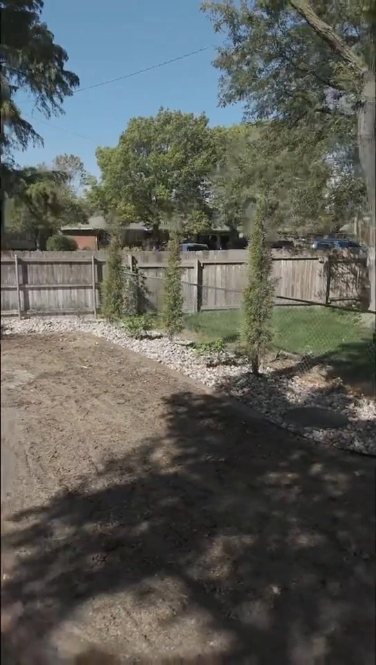 A row of young columnar evergreens set in river rock along the back fence of the Brown residence, with cleared ground in front of them.