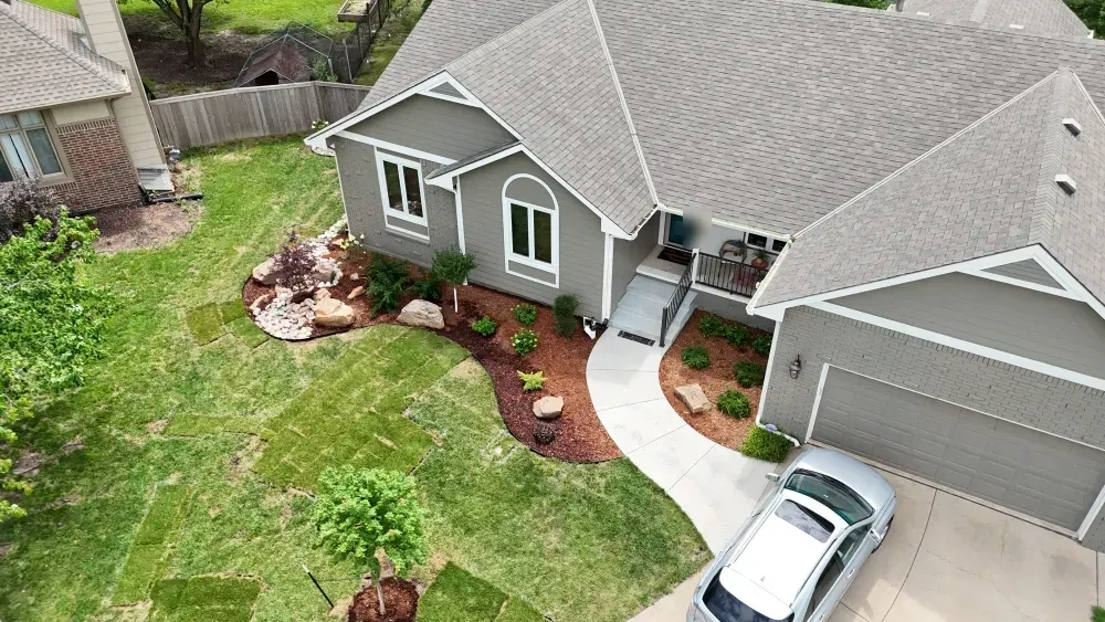 The Graber front bed from above the drive: mulch, boulders and new shrubs along the house, the walk curving to the porch, and new sod around a staked young tree.
