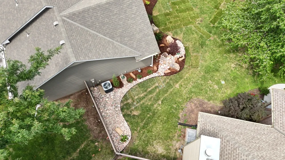 An aerial view of a cobble drainage run curving between a house and its lawn, edged in mulch and set with boulders and new planting.