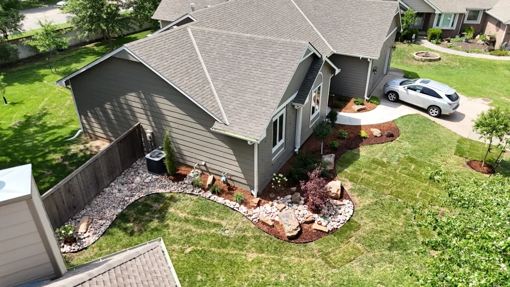 The side of a house from above: a curving river rock border set with boulders runs along the wall and around the air conditioner, between a wood fence and new sod.