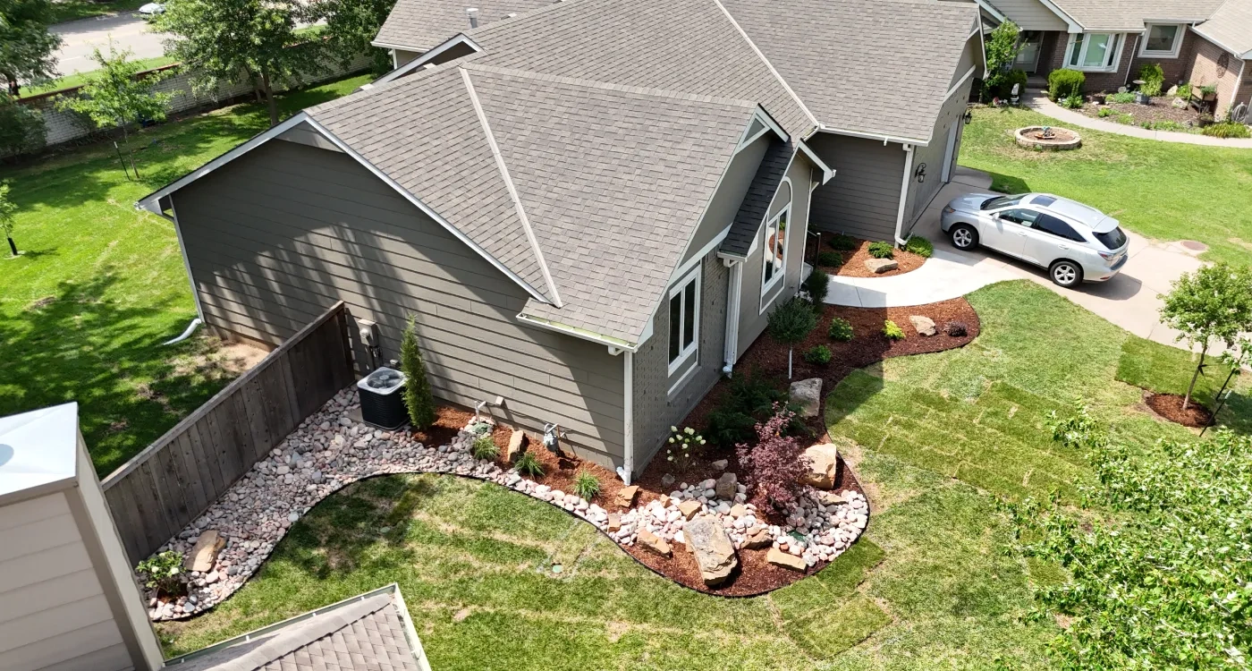 The Graber residence from above: river-rock beds curving around the house past a cedar fence, fresh sod squares knitting into the lawn and the drive at right.