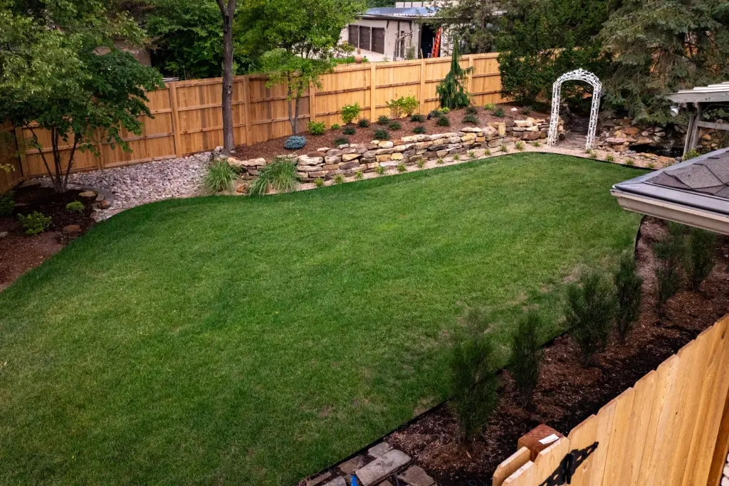 The Grainger back garden from the deck: new sod ringed by mulched beds, the boulder retaining wall along the cedar fence and the white arbour in the corner.