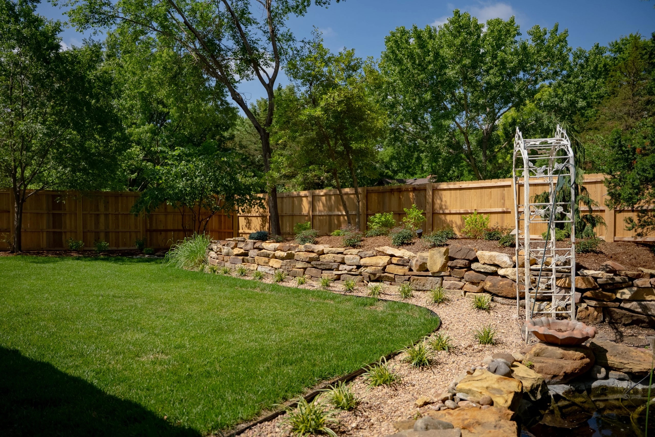 The back garden's dry-stacked stone wall under the cedar fence, the white arbour at its corner and the new lawn cut in below.