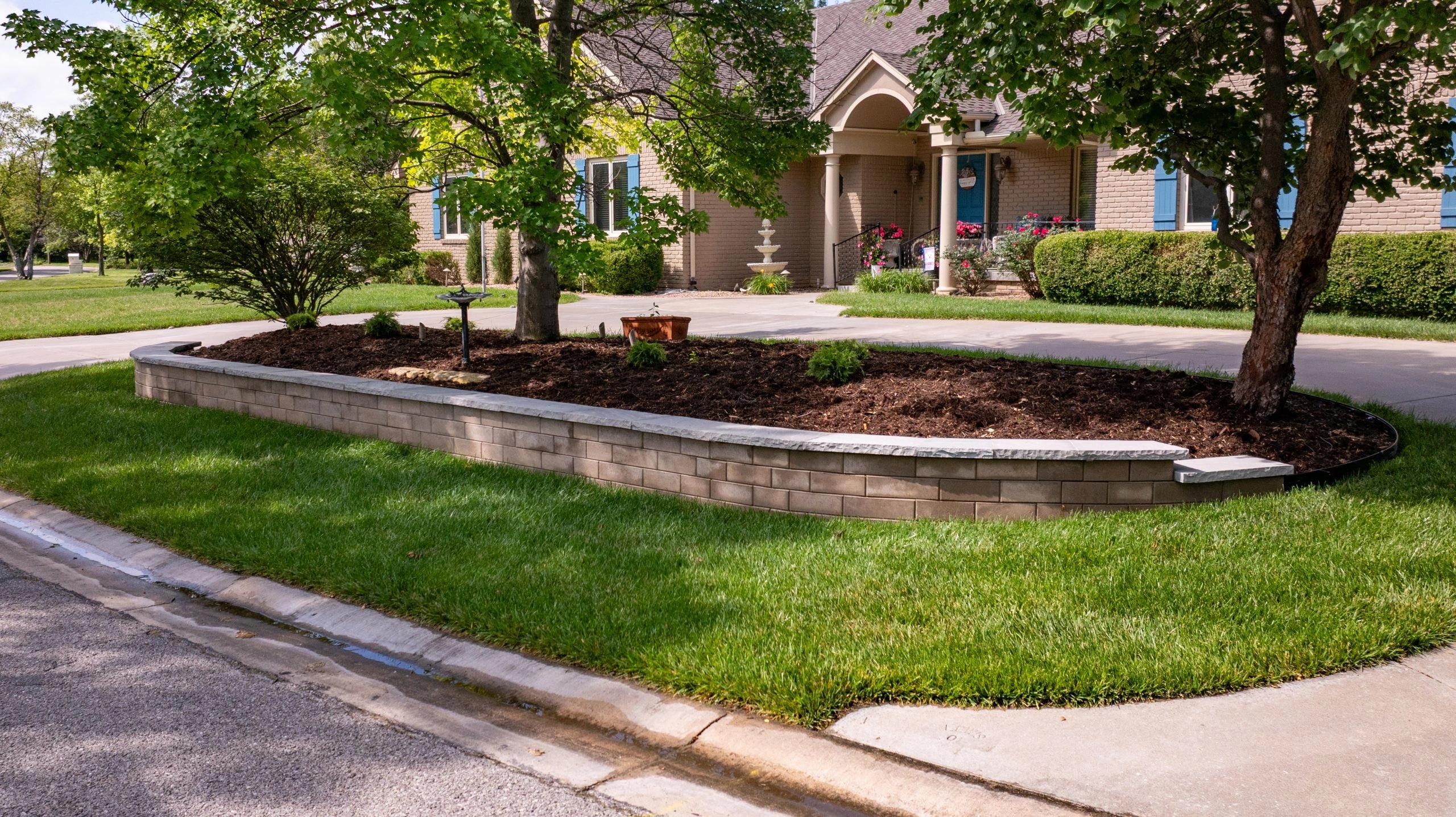 The front corner bed: a curved block retaining wall with a stone cap holding a mulched island of trees and shrubs, the drive and house behind it.