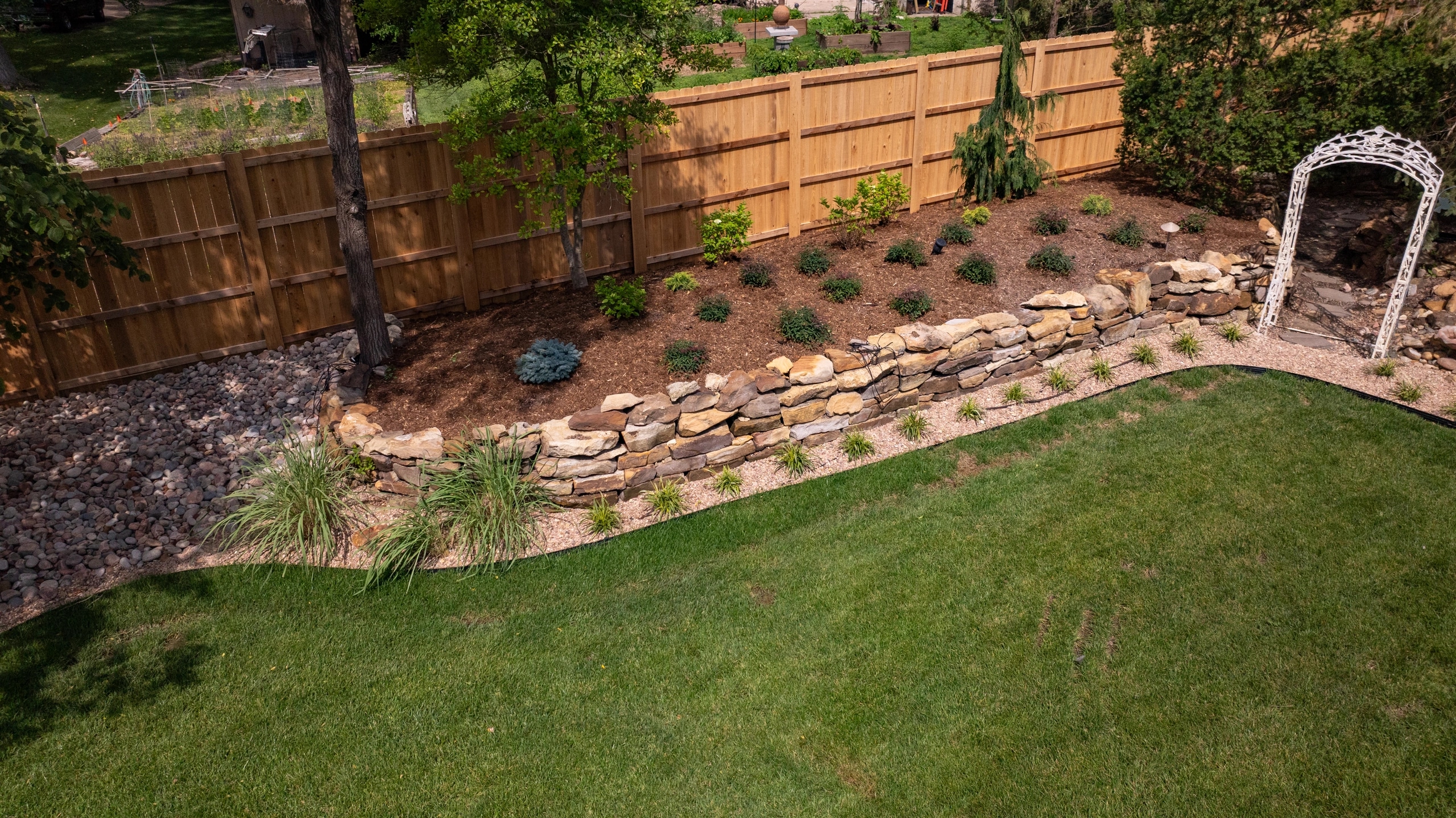 From above: a black edge curves along a level lawn, holding a strip of pale gravel and grasses at the foot of a dry-stacked stone wall and a mulched bed.