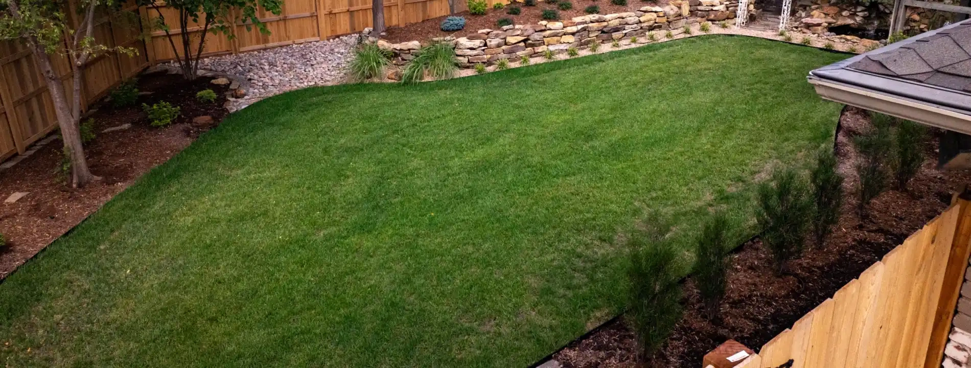 A shaped lawn edged in aluminium, with a pale gravel strip running along the fence line and a dry-stacked stone wall holding the planted bank behind it.