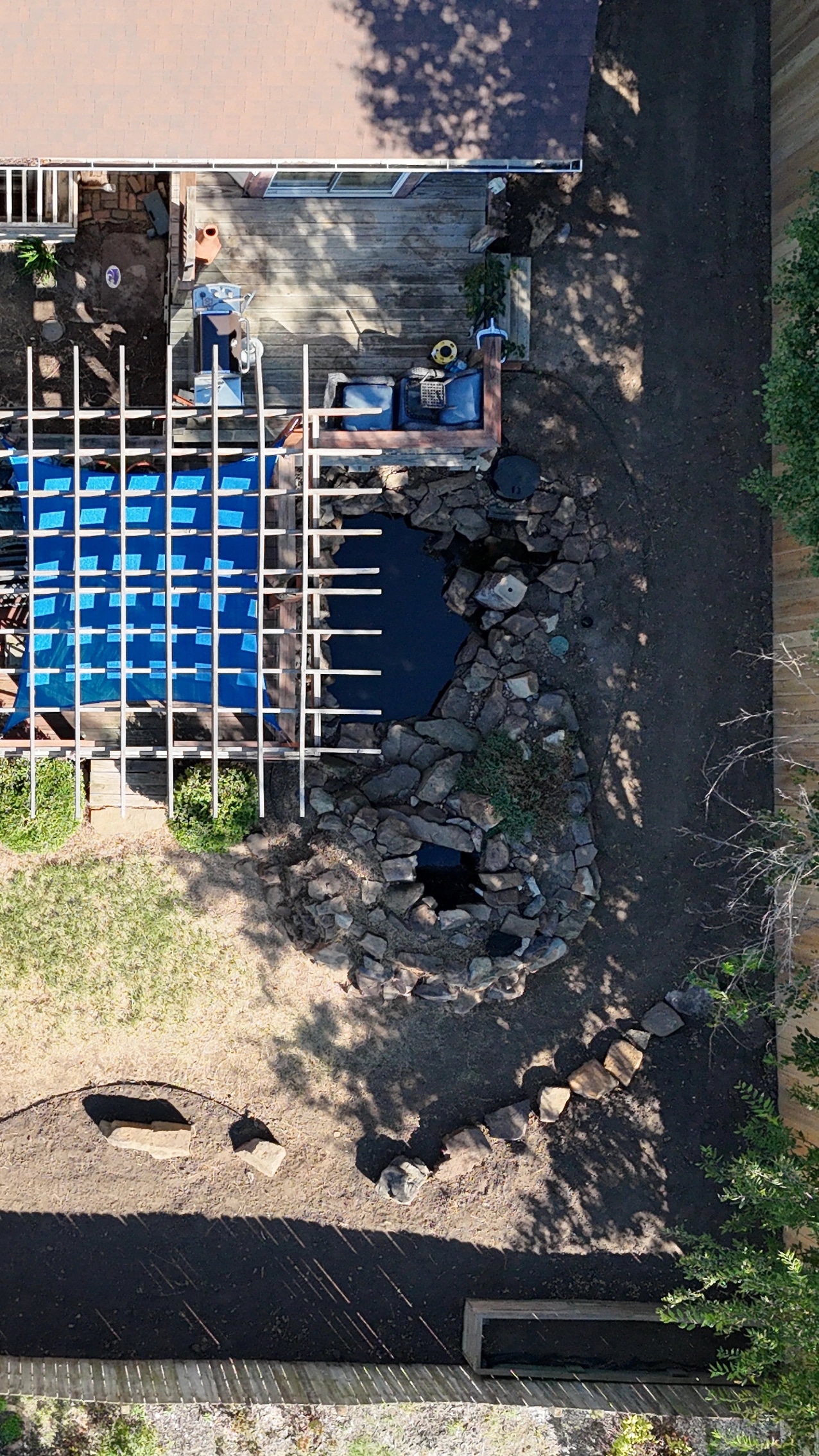 Straight down on the finished yard: the pergola deck and the stone-edged pond, a curving line of stone across the cleared ground, and the fence.
