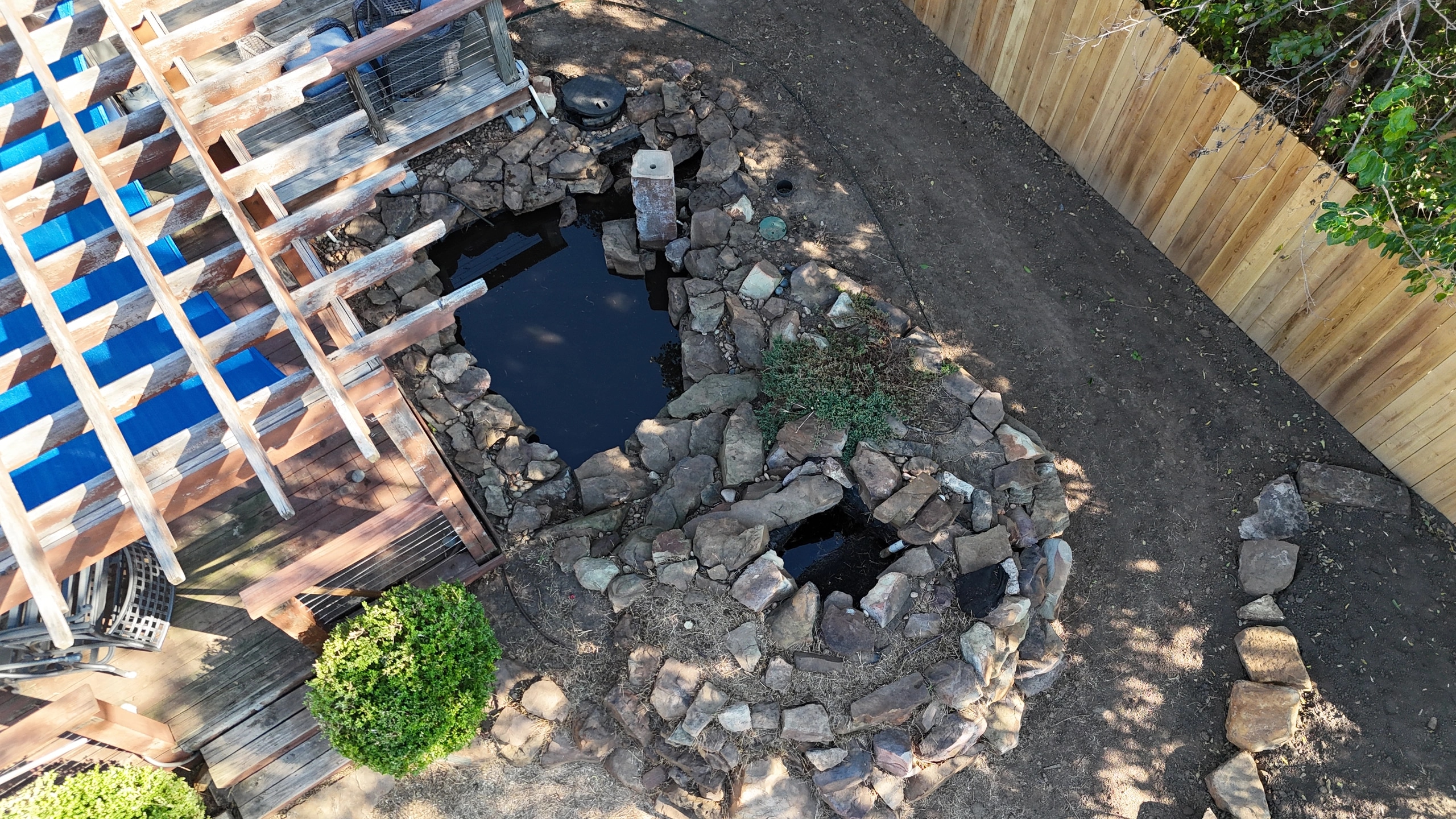 The same pond after the cleanup: open water inside a restacked stone edge, the pergola deck beside it and cleared ground running to the new fence.