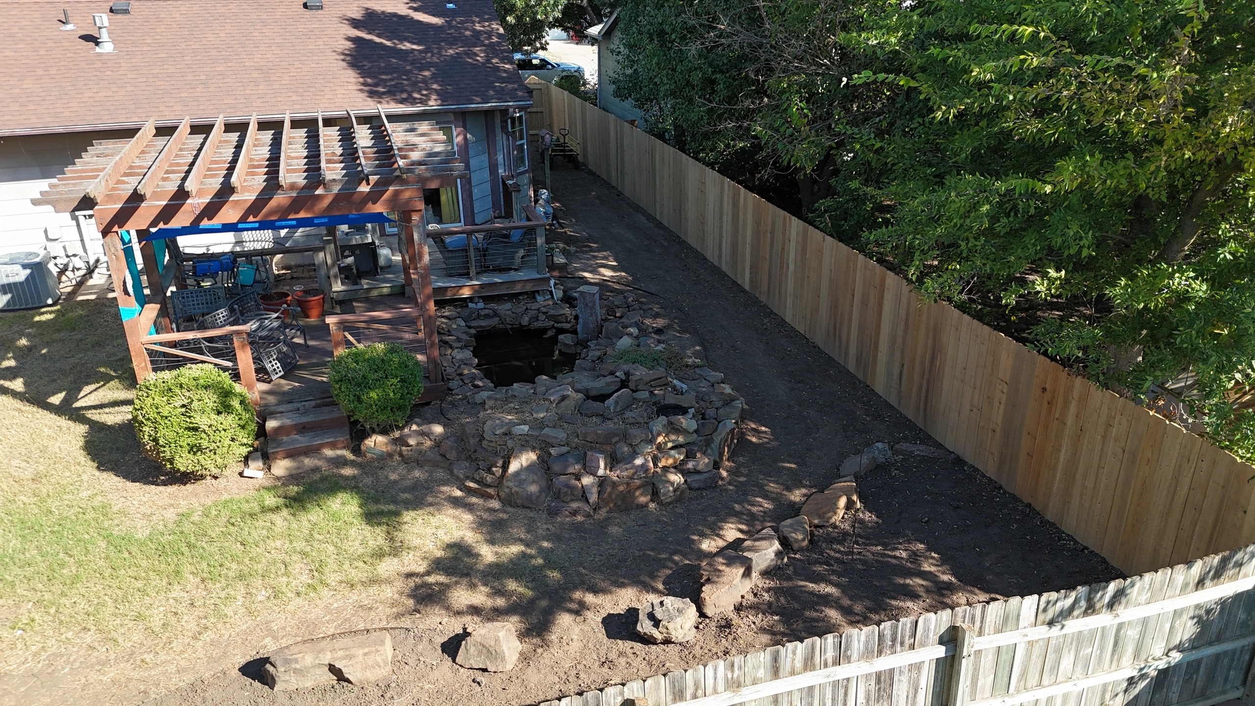 The cleared back yard from the far corner: bare ground along a new board fence, the pond’s stone edge restacked, and trimmed shrubs at the pergola deck steps.