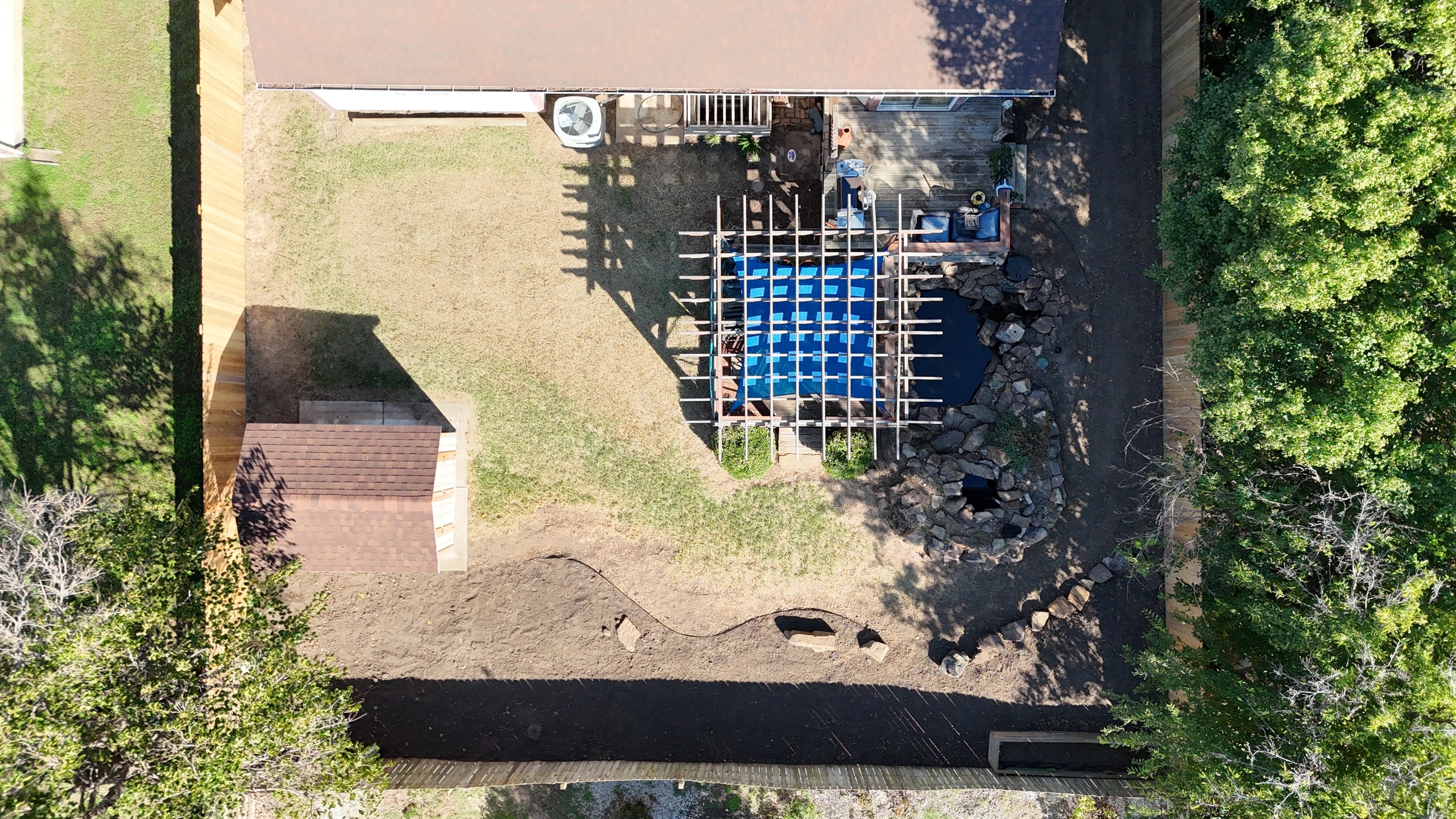 The same yard straight down after the cleanup: cleared ground along the fence lines, the stone-edged pond beside the pergola, and stone set in a curving line.