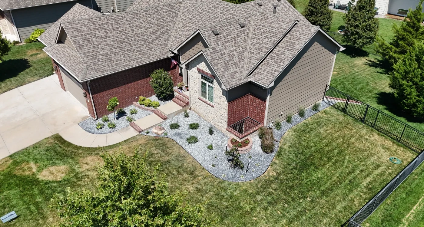 A finished front landscape from above: gravel beds of new shrubs curving around a brick and stone house, a concrete entry walk, and mown lawn inside a black iron fence.