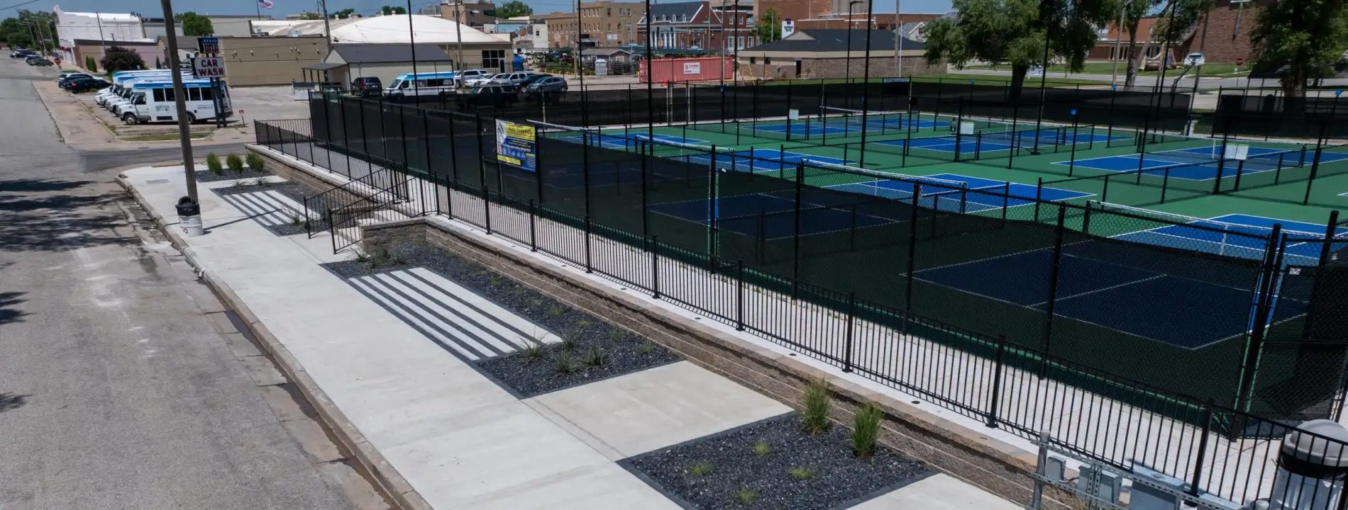 The finished entrance to the McPherson pickleball courts seen from the road, with a black iron fence along the new concrete walk and the painted courts behind it.